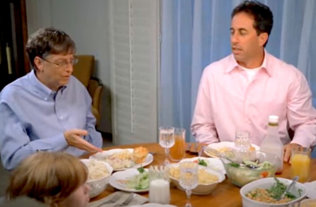 A still from a Microsoft ad shows Bill Gates and Jerry Seinfeld eating dinner with a family.