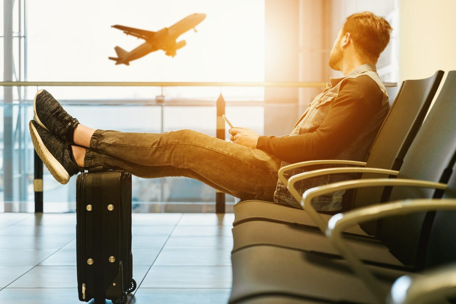 A man sits in an airport terminal with his feet resting on his suitcase and an iPhone with eSIM for international data in his hand. A plane is taking off outside the window.
