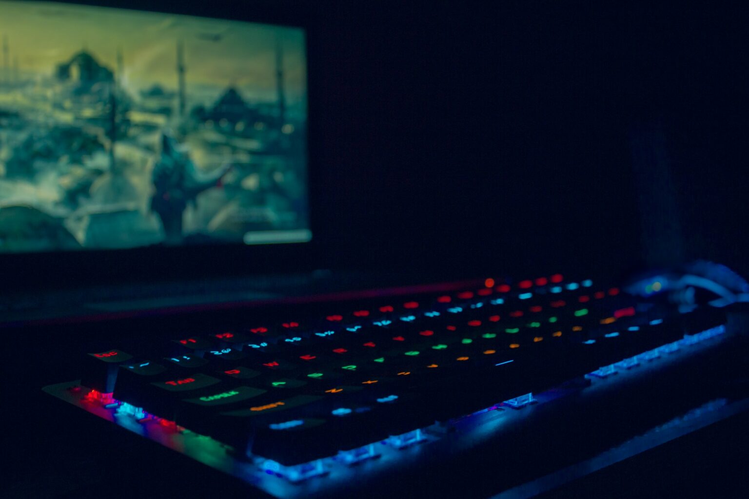 A gaming PC keyboard with colorful RGB lighting sits in front of a computer monitor in a darkened room.