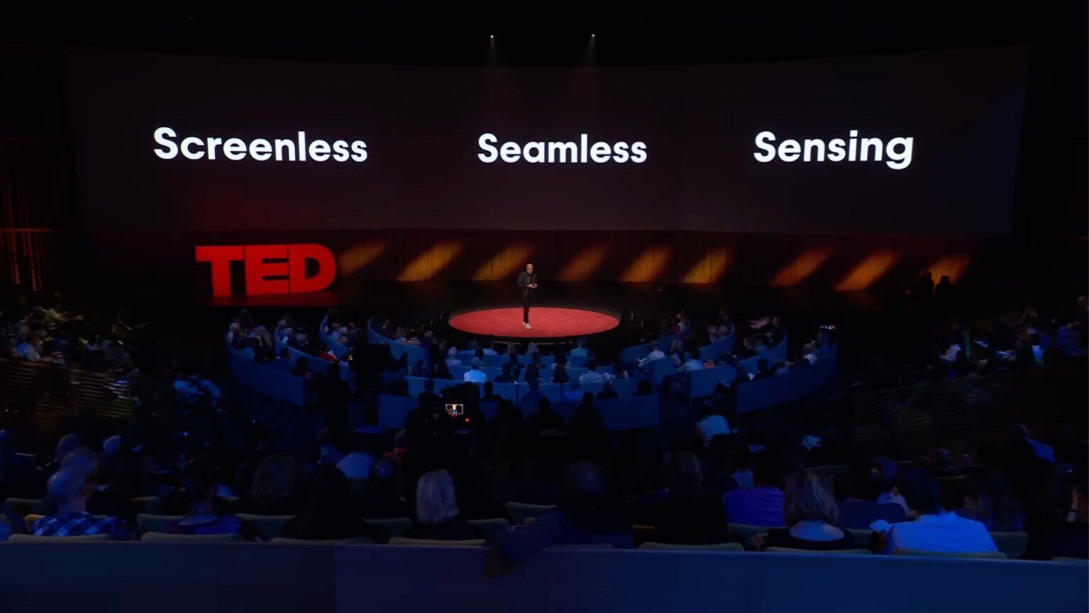 Imran Chaudhri standing on stage at a TED Talk with the phrase “Screenless Seamless Sensing” written on a slide behind him