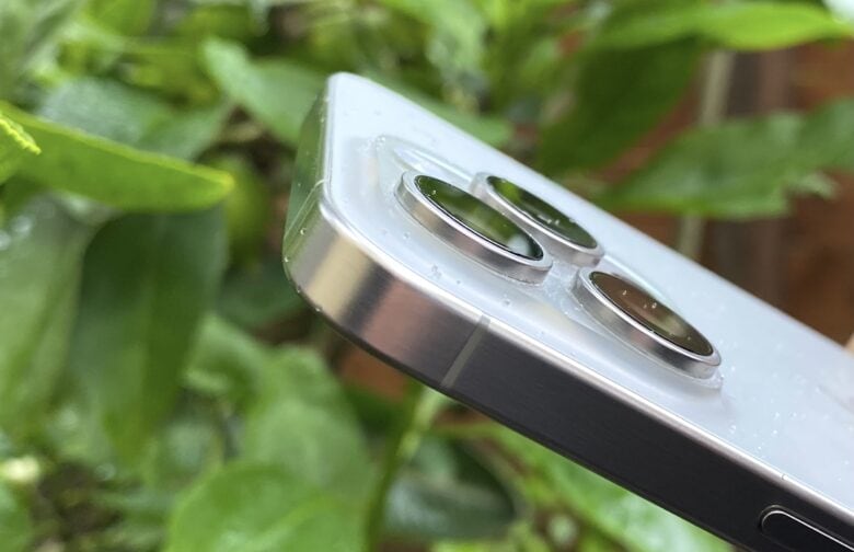 iPhone 15 Pro Max in natural titanium with some water droplets on it, shown in front of a green plant.