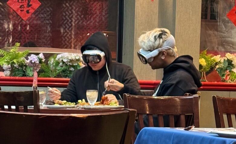 Two people at a restaurant eating while wearing Vision Pro headsets.