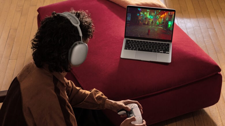 Man playing Stray on his MacBook Air (which isn’t plugged in) wearing AirPods Max and holding a PlayStation 5 controller