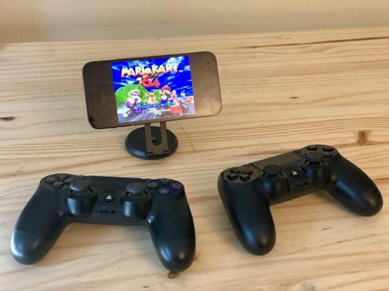 iPhone sitting on SwitchEasy stand with two PS4 controllers playing Mario Kart 64 on Delta game emulator for iPhone