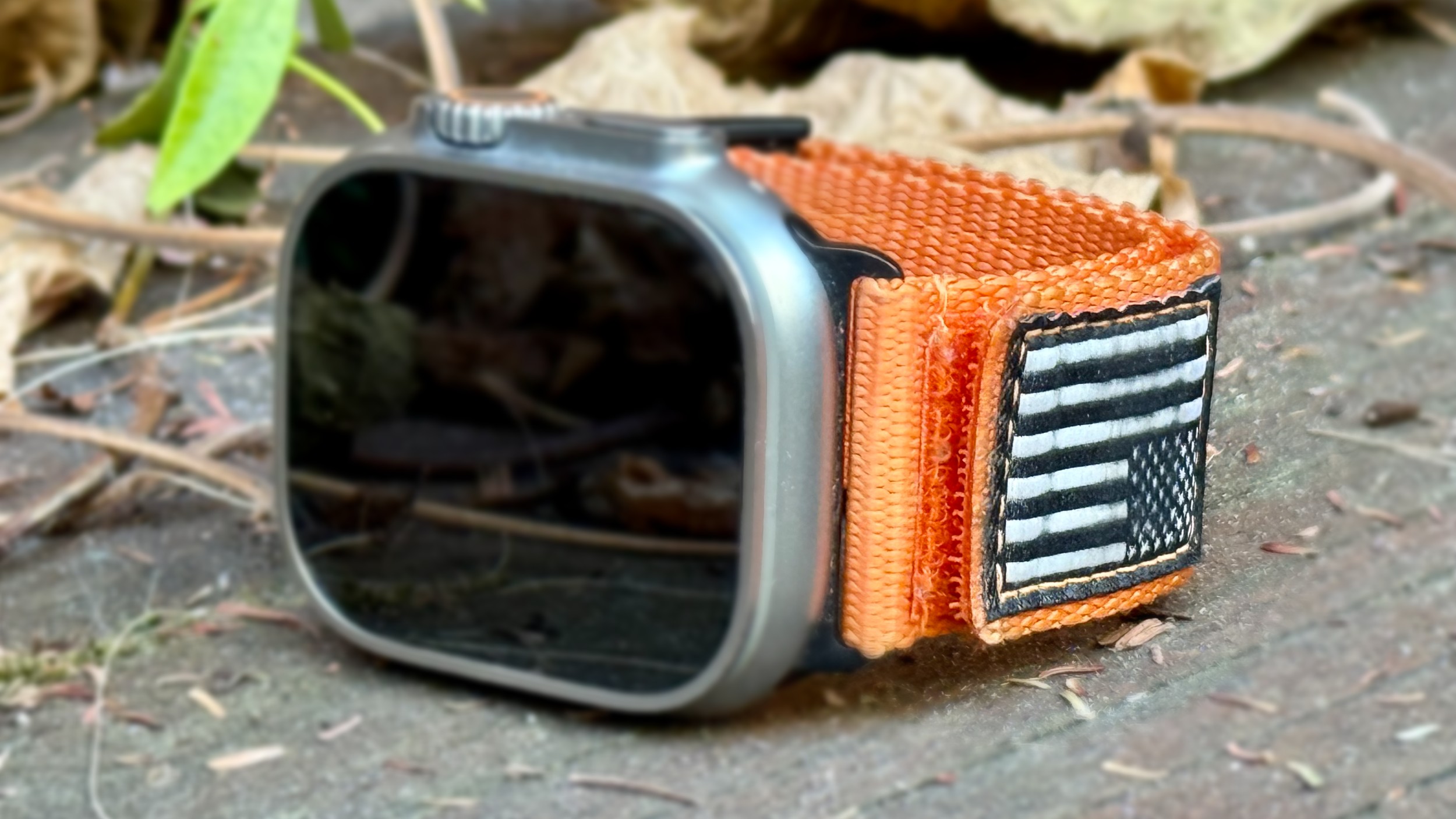 American flag Apple Watch Ultra band orange Orange nylon band with American flag, attached to an Apple Watch Ultra and sitting on a bench in an outdoor setting.