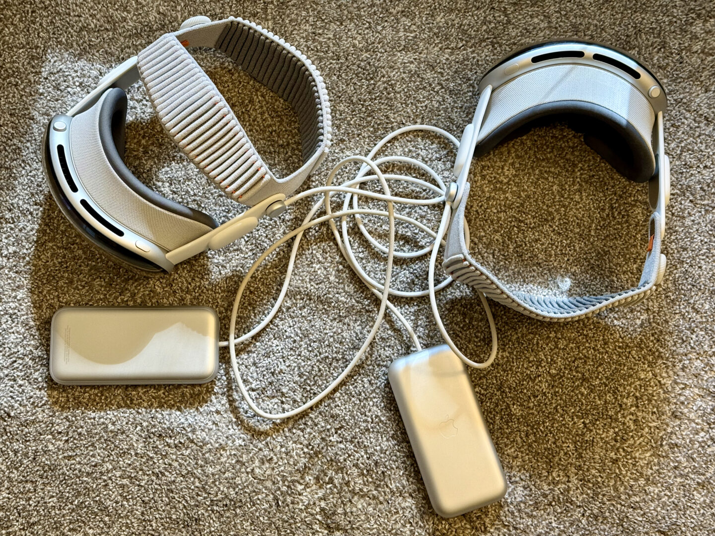 M5 M2 Vision Pro Batteries Two Vision Pros sitting on a gray carpet, their batteries and cables in a tangled mess.