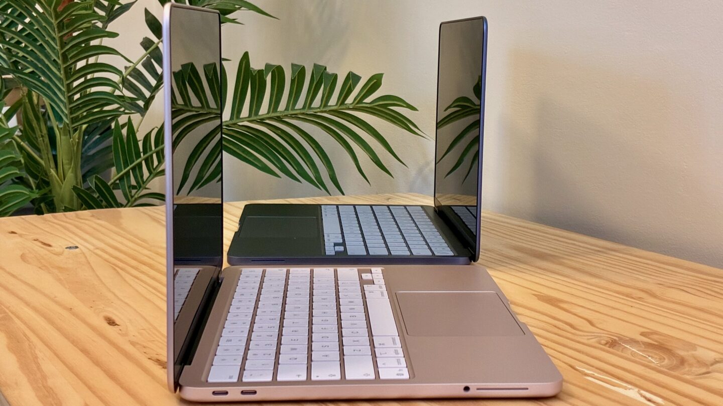 MacBook Neo in Blush and Indigo, lined up, sitting on a wood table in front of a fake tree.