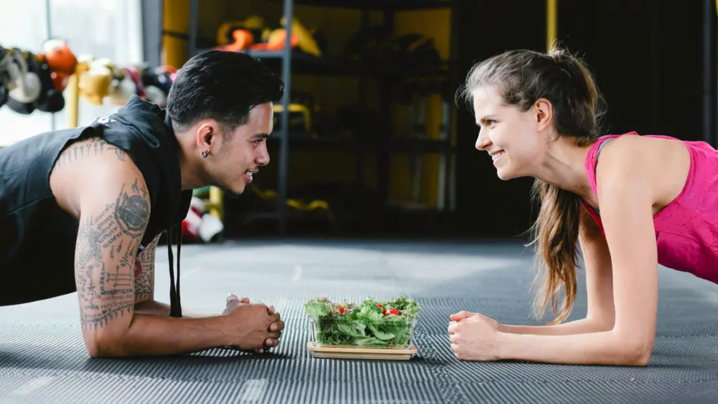 Photo of a man and a woman doing planks in a gym with a salad between them, used to illustrate a post about NutriAI nutrition and workout app