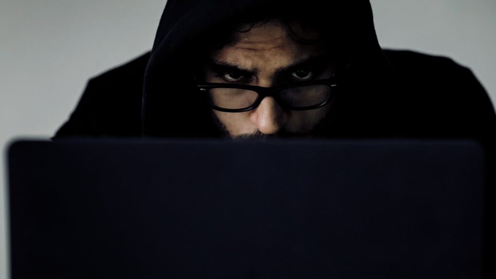A photo of a man wearing a black hoodie, peering ominously at a laptop screen